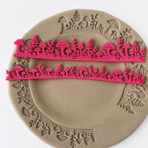 May include: Two long, thin, bright pink strips featuring a detailed cutout pattern of various mushrooms and fern-like foliage. These strips are positioned horizontally across a circular, light brown clay plate that has matching mushroom and fern impressions.
