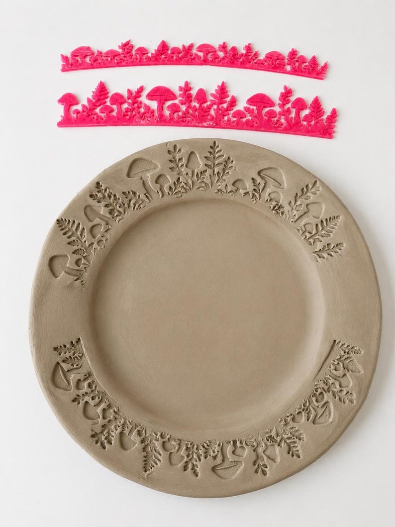 May include: Two bright pink flexible silicone stamps featuring intricate mushroom and fern patterns, positioned above a circular gray clay plate that displays the same embossed botanical designs pressed into its outer rim surface.