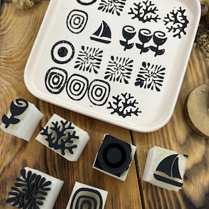 Op de afbeelding: Een vierkant gebroken wit keramieken dienblad met een raster van zwarte gestempelde ontwerpen, waaronder cirkels, koraalachtige vormen, een zeilboot en bloemen. Zeven individuele vierkante rubberen stempels met bijpassende zwarte ontwerpen liggen eromheen.