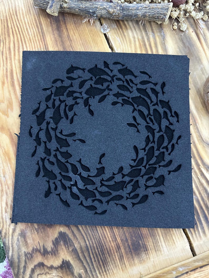 May include: A square, dark gray coaster featuring a school of fish arranged in a circular pattern. The fish are cut out of the coaster, creating a negative space design. The coaster is made of a textured material.