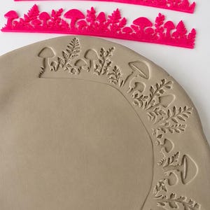 May include: Two bright pink plastic stamps featuring a repeating pattern of mushrooms and ferns, positioned above a circular piece of light gray clay that has been impressed with the same mushroom and fern design along its outer edge.