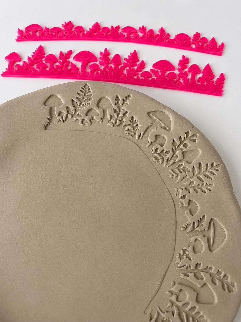 May include: Two bright pink plastic stamps featuring a repeating pattern of mushrooms and ferns, positioned above a circular piece of light gray clay that has been impressed with the same mushroom and fern design along its outer edge.