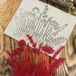 May include: A beige clay mould with an embossed floral design, featuring various wildflowers and grasses. A separate, vibrant red floral piece is placed below the mould, showcasing the potential result. The items are set on a wooden surface.