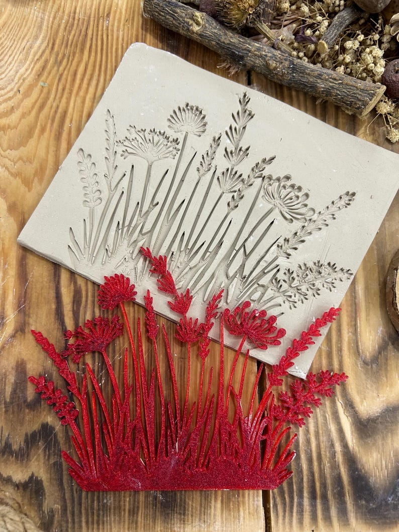 May include: A beige clay mould with an embossed floral design, featuring various wildflowers and grasses. A separate, vibrant red floral piece is placed below the mould, showcasing the potential result. The items are set on a wooden surface.