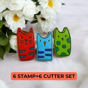 May include: A set of three colorful cat-shaped stamps and cutters. The stamps are red, blue, and green, with decorative stripes and dots. The text "6 STAMP+6 CUTTER SET" is displayed in an orange rectangle.