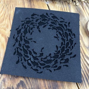 May include: A square, dark blue piece of fabric with a circular design of cutout fish. The fish are arranged in a ring, creating a decorative pattern. The fabric has a slightly textured appearance.