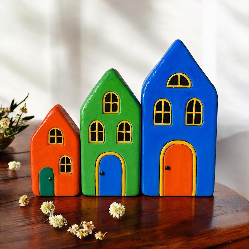 May include: Three colorful wooden house figurines. The houses are painted in bright orange, green, and blue, each with yellow-framed windows and doors. The houses are arranged in a row, with the smallest on the left and the largest on the right.