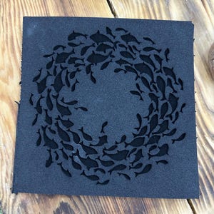 May include: A square, dark gray coaster featuring a school of fish arranged in a circular pattern. The fish are cut out of the coaster, creating a negative space design. The coaster is made of a textured material.