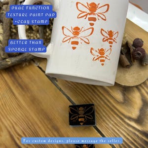 Può includere: Una tazza di ceramica bianca con disegni di api arancioni. La tazza presenta il testo "DUAL FUNCTION TEXTURE PAINT PAD + CLAY STAMP" e "BETTER THAN SPONGE STAMP". È visibile anche un piccolo timbro a forma di ape. Il testo "For custom designs, please message the seller" è in basso.