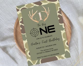 May include: A birthday invitation with a camouflage pattern border. The invitation features the text "The Big ONE" with a target graphic and antler accents. It reads "Hunter's First Birthday" with event details.
