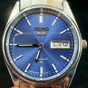 May include: A stainless steel Seiko 5 automatic watch with a blue dial. The watch face features silver hour markers and hands, a day-date display, and the text "SEIKO 5" and "Tomatic". The watch has a silver strap.