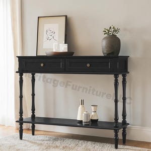 May include: A black console table with two drawers and brass knobs. The table has turned legs and a lower shelf. Decorative items on the table include a framed print, candles, and vases. A vase with greenery sits on the top.
