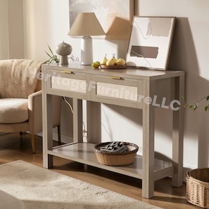 May include: A light gray wooden console table with two drawers and a lower shelf. The drawer fronts have woven cane inserts and gold-colored handles. A woven basket sits on the shelf. A lamp, artwork, and decorative items are on top.