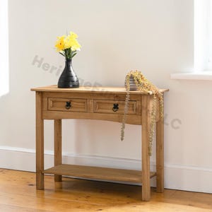 May include: A light brown wooden console table with two drawers and a lower shelf. The table has black hardware and is decorated with a black vase of yellow flowers and a small decorative plant. The table is 48 inches long.