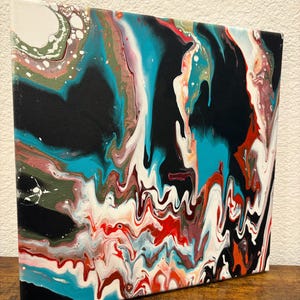 May include: Abstract acrylic painting on canvas with a fluid pour technique. The artwork features a mix of colors including black, teal, red, white, and brown, creating a dynamic and textured visual effect. The canvas is mounted on a wooden frame.