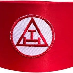 Può includere: Un fez rosso vivo con un emblema circolare bianco con un triangolo rosso e un design a barra orizzontale. Il cappello è cilindrico e realizzato in un materiale liscio, possibilmente satinato.