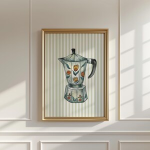 May include: Framed print of a vintage-style coffee maker. The coffee pot is light blue with orange and yellow floral accents. It has a black handle and sits against a striped background in a gold frame. The artwork is displayed on a white wall.