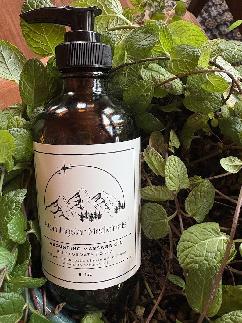 Puede incluir: Una botella de vidrio marr&oacute;n con un dispensador de bomba negro, etiquetada como "Morningstar Medicinals Grounding Massage Oil". La etiqueta presenta un dise&ntilde;o de monta&ntilde;a y &aacute;rboles. La botella contiene 237 ml de aceite y est&aacute; rodeada de plantas verdes.