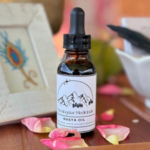 Puede incluir: Una botella de vidrio marrón de aceite Nasya con un cuentagotas negro, etiquetada como "Morningsar Medicinals". La etiqueta presenta un diseño de montaña y árboles. Pétalos de rosa rosas y blancos están esparcidos alrededor de la botella sobre una bandeja de madera. La botella contiene 30 ml de aceite.