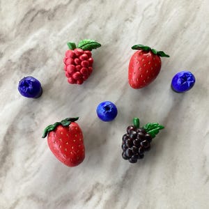 May include: A collection of handcrafted berry-shaped objects, including strawberries, raspberries, blueberries, and grapes. The berries are painted in vibrant red, purple, and blue hues, with green leaves. The arrangement is set against a light marble surface.