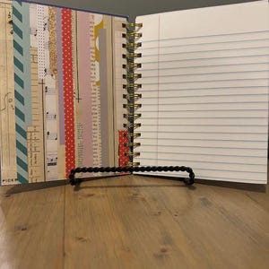 Pode incluir: Di&aacute;rio aberto com espiral, com p&aacute;ginas pautadas e detalhes decorativos em papel. O lado esquerdo apresenta tiras de papel estampado, incluindo notas musicais e bolinhas. O di&aacute;rio &eacute; mantido aberto por um suporte preto com contas, sobre uma superf&iacute;cie de madeira.