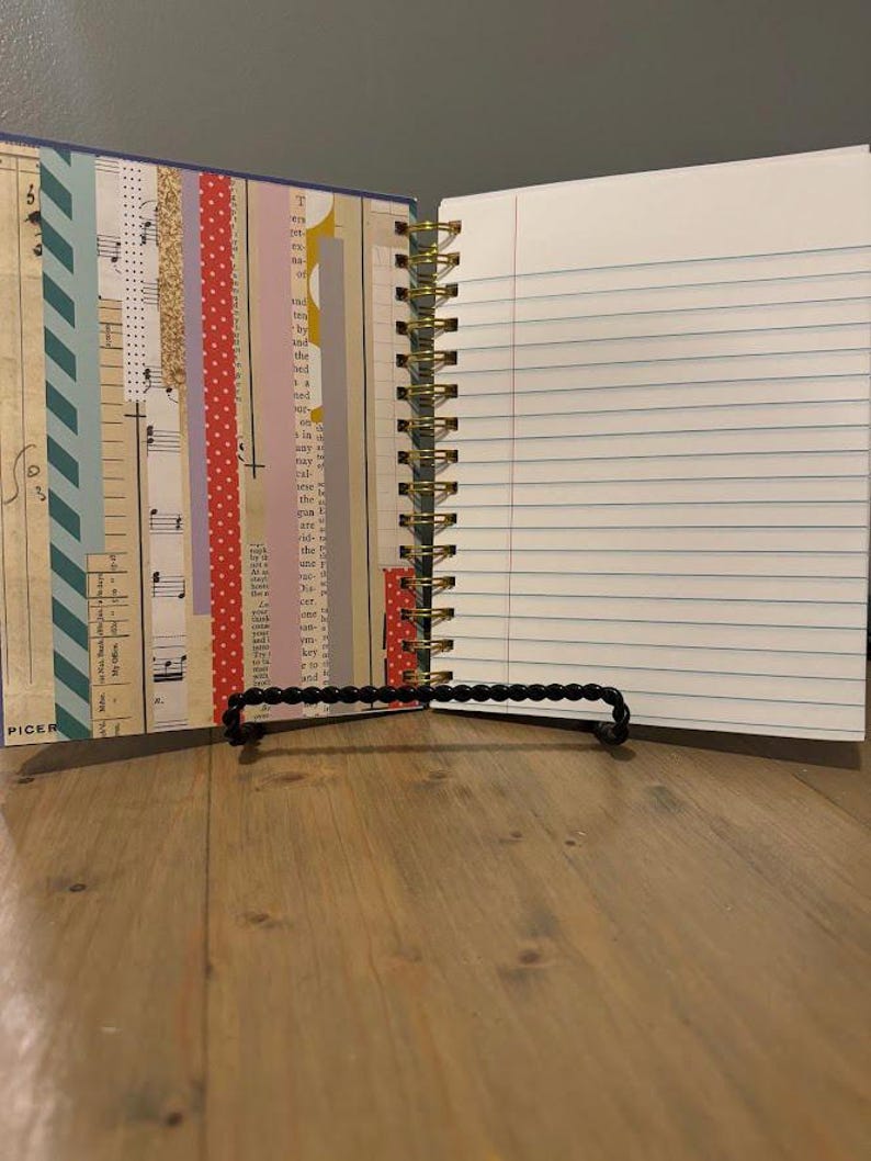 Pode incluir: Di&aacute;rio aberto com espiral, com p&aacute;ginas pautadas e detalhes decorativos em papel. O lado esquerdo apresenta tiras de papel estampado, incluindo notas musicais e bolinhas. O di&aacute;rio &eacute; mantido aberto por um suporte preto com contas, sobre uma superf&iacute;cie de madeira.