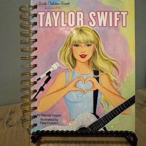 Pode incluir: Um livro com espiral intitulado "Taylor Swift" da s&eacute;rie biogr&aacute;fica "Little Golden Book". A capa apresenta uma ilustra&ccedil;&atilde;o de Taylor Swift segurando uma guitarra e formando um cora&ccedil;&atilde;o com as m&atilde;os. O livro &eacute; exibido em um suporte de metal preto.
