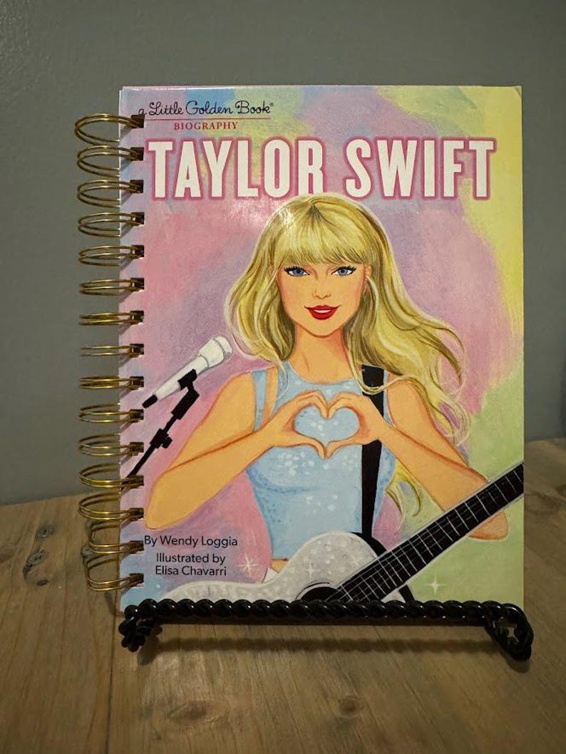 Pode incluir: Um livro com espiral intitulado "Taylor Swift" da s&eacute;rie biogr&aacute;fica "Little Golden Book". A capa apresenta uma ilustra&ccedil;&atilde;o de Taylor Swift segurando uma guitarra e formando um cora&ccedil;&atilde;o com as m&atilde;os. O livro &eacute; exibido em um suporte de metal preto.