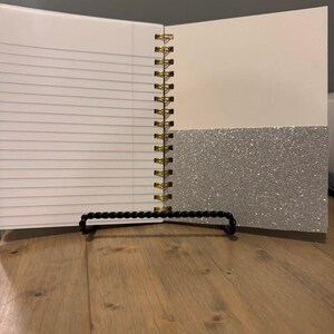 Pode incluir: Caderno aberto com p&aacute;ginas pautadas e encaderna&ccedil;&atilde;o em espiral dourada. O lado direito tem uma p&aacute;gina bege clara com um bolso de glitter prateado. O caderno &eacute; apoiado em um suporte de contas pretas.