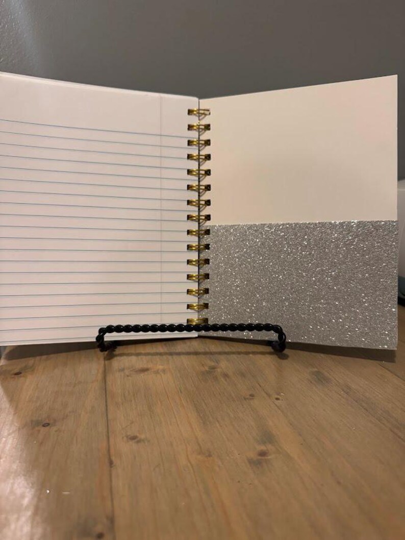 Pode incluir: Caderno aberto com p&aacute;ginas pautadas e encaderna&ccedil;&atilde;o em espiral dourada. O lado direito tem uma p&aacute;gina bege clara com um bolso de glitter prateado. O caderno &eacute; apoiado em um suporte de contas pretas.
