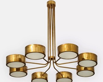 May include: A gold-toned chandelier with eight cylindrical light fixtures. Each fixture has a gold exterior and a white interior. The chandelier is suspended from the ceiling by multiple gold rods.