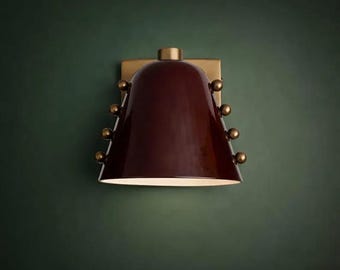 May include: A wall-mounted light fixture with a burgundy shade and brass accents. The shade is bell-shaped and has a glossy finish. Small brass spheres adorn the sides. The fixture is mounted on a square brass plate.