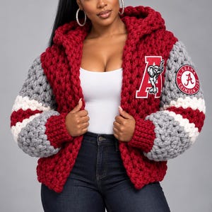 May include: A chunky knit hooded cardigan in deep crimson red with grey and white striped sleeves. The front features a large white letter A patch with an elephant graphic, and the left sleeve displays a circular Alabama Crimson Tide logo patch.