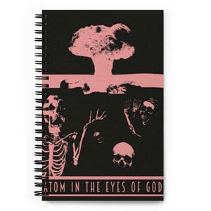 May include: Spiral-bound notebook with a black cover featuring a pink mushroom cloud illustration. Below the cloud are skeletal figures and skulls. The text "ATOM IN THE EYES OF GOD" is printed at the bottom.