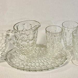 Puede incluir: Un juego de agua de vidrio transparente con una jarra, seis vasos y una bandeja de servir. La jarra tiene un asa y un pico. Los vasos son de diferentes tamaños. El juego tiene un patrón texturizado, similar a burbujas. La bandeja es redonda con un borde festoneado.