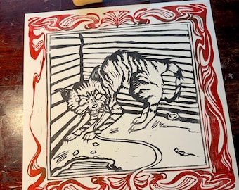 May include: A black and white linocut print of a cat with tiger-like stripes, framed in red decorative borders. The cat is in a dynamic pose, possibly pouncing. A carving tool rests above the print on a wooden surface.