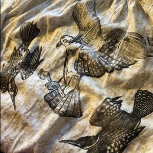 May include: Cream-colored fabric with a repeating floral pattern. The fabric is printed with detailed black and gray illustrations of birds in various poses, including a bird of prey with outstretched wings. The overall theme is nature and wildlife.