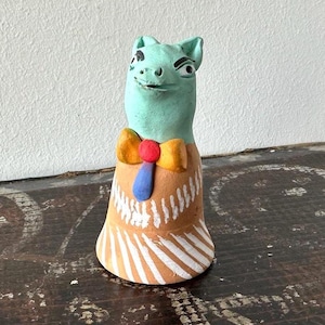 May include: A whimsical, hand-painted ceramic figurine of a creature with a turquoise head and a brown body. It features a yellow bow tie with a red center and a blue tie. The base is decorated with white stripes.