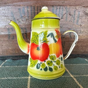 May include: Vintage enamelware coffee pot with a bright green lid, spout, and handle. The body is decorated with a colorful fruit and vegetable design, including red apples, yellow pears, and green leaves. The pot has a white handle and a small knob on the lid.