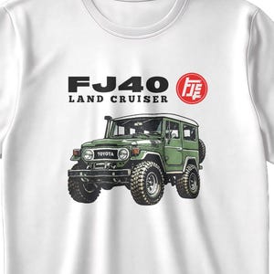 以下が含まれることがあります： 緑色のFJ40ランドクルーザーのグラフィックが特徴の白いTシャツ。「FJ40 LAND CRUISER」の黒い文字と赤い日本の文字がデザインに含まれています。トヨタのロゴも表示されています。シャツは柔らかい素材で作られています。
