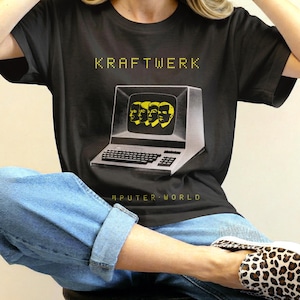 Peut inclure: Un t-shirt noir à manches courtes avec un graphique représentant un écran d'ordinateur vintage affichant quatre visages jaunes. Le texte KRAFTWERK figure au-dessus et COMPUTER WORLD est imprimé en dessous dans une police pixelisée jaune.