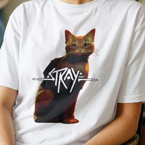 May include: A white short-sleeved t-shirt featuring a graphic illustration of a brown and black cat sitting upright. The word STRAY is printed in a stylised white font across the centre of the cat's body, with horizontal lines extending outwards.