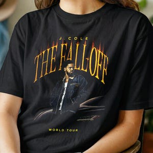 May include: A black short-sleeve t-shirt featuring a graphic print of J. Cole in a denim jacket. The text reads J. COLE THE FALL OFF WORLD TOUR in a yellow, flame-like font. The design includes a dark car silhouette behind the figure.