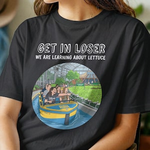 Pode incluir: Uma t-shirt preta com o texto branco GET IN LOSER WE ARE LEARNING ABOUT LETTUCE sobre um gráfico circular de um passeio de barco amarelo e azul através de uma estufa cheia de filas de alfaces verdes e pessoas.
