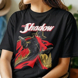 Könnte beinhalten: Ein schwarzes Kurzarm-T-Shirt mit einer Grafik eines Charakters in schwarzem Hut, Trenchcoat und rotem Schal, der zwei Handfeuerwaffen hält. Der Text THE SHADOW ist in Weiß und Rot über der Figur gedruckt, mit einer gelben Skyline darunter.