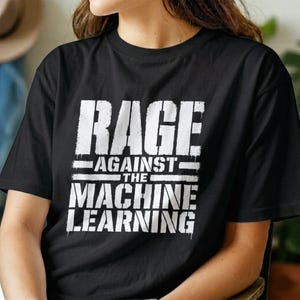 Peut inclure: Un t-shirt noir à manches courtes avec un texte blanc au style vieilli sur le devant qui indique RAGE AGAINST THE MACHINE LEARNING. Le motif graphique est centré sur la poitrine et utilise une police grasse avec un aspect texturé et usé.