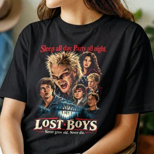 Può includere: Una t-shirt nera con una stampa grafica del film The Lost Boys. Il design include il testo Sleep all day. Party all night. in alto, un collage di personaggi tratti dal film e il titolo LOST BOYS stampato nella parte inferiore del disegno.