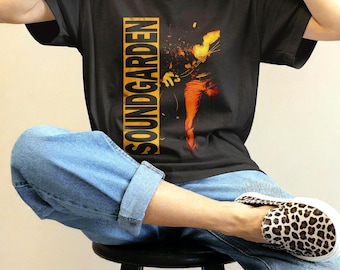 May include: A black short-sleeve t-shirt featuring the word SOUNDGARDEN printed vertically in yellow within a rectangular border, alongside a graphic illustration of a figure in orange and yellow tones, paired with blue denim jeans and leopard print shoes.