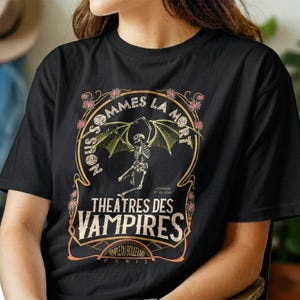 Puede incluir: Una camiseta negra con un estampado gráfico de un esqueleto con alas de murciélago. El diseño incluye el texto "NOUS SOMMES LA MORT", "THEATRES DES VAMPIRES", "VENDREDI 3 DU SOIR" y "TEMPLE DU BOULEVARD PARIS" enmarcado por elementos florales.