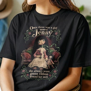 Puede incluir: Una camiseta negra con una ilustración gráfica de una niña de pelo oscuro sentada en un sofá, sosteniendo un ramo de flores y una muñeca pequeña. El texto dice: Once there was a girl named Jenny, she always wore green ribbon around her neck.